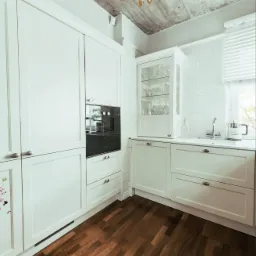 Biała kuchnia z drewnianą podłogą i surowym betonowym sufitem, oświetlona złotym żyrandolem; widoczne białe płytki na ścianie i zabudowa kuchenna z piekarnikiem i szklaną witryną.