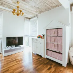 Jasne wnętrze pokoju dziecięcego z drewnianą podłogą, białymi meblami z różowymi koszami, telewizorem i workiem do siedzenia w gwiazdki, sufit betonowy z widoczną instalacją elektryczną i złotym...