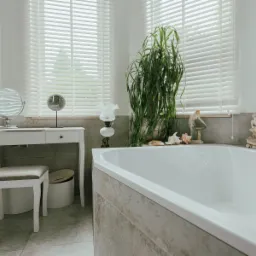 Jasna łazienka z wanną, toaletką z dwoma lustrami, wysoką rośliną doniczkową oraz dekoracjami w postaci figurek i muszli, oświetlona światłem dziennym wpadającym przez żaluzje.