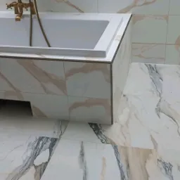 Wykończona łazienka z wanną obudowaną płytkami imitującymi marmur, złota bateria, płytki podłogowe i ścienne z podobnym wzorem.