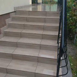 Zewnętrzne schody wejściowe wyłożone płytkami imitującymi drewno, z czarną metalową barierką, po deszczu.