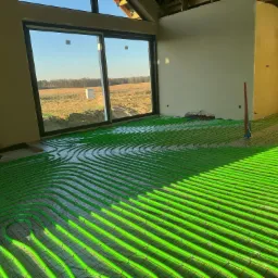 Zielone rury ogrzewania podłogowego ułożone w surowym wnętrzu z dużym oknem, przez które wpada światło słoneczne. Widoczne elementy konstrukcji dachu.