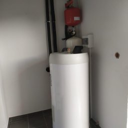 Michał Miklaszewski SUNLIT - Biały, cylindryczny zbiornik akumulacyjny OSO z czarnymi rurami instalacji grzewczej i czerwonym naczyniem wzbiorczym w rogu pomieszczenia z szarą podłogą.