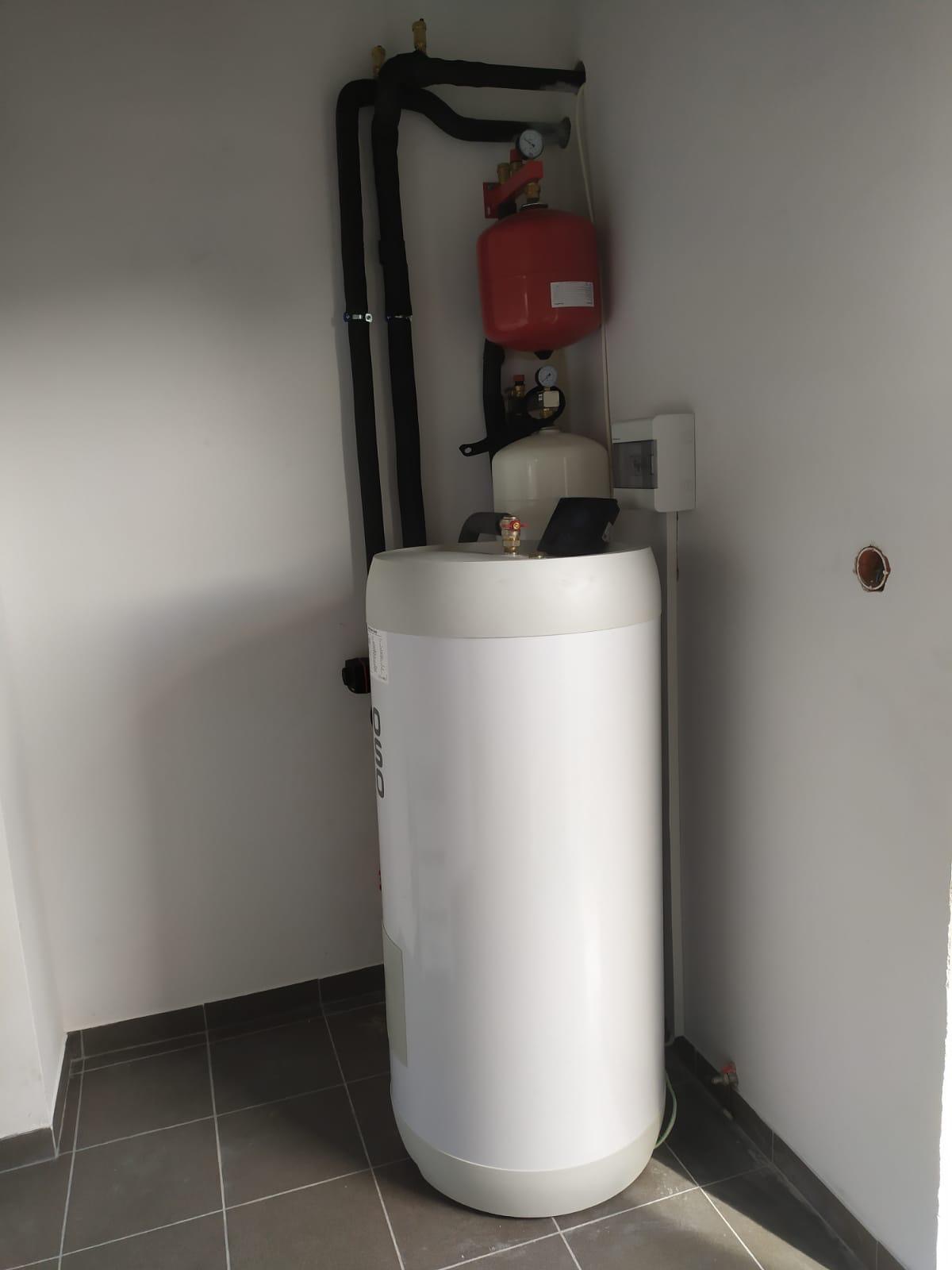 Biały, cylindryczny zbiornik akumulacyjny OSO z czarnymi rurami instalacji grzewczej i czerwonym naczyniem wzbiorczym w rogu pomieszczenia z szarą podłogą.