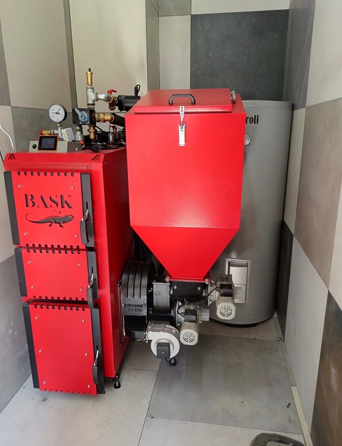 Czerwony piec na pellet marki Baski z podajnikiem, model Technix SV-300, obok szary zasobnik na wodę w kotłowni z szaro-białymi płytkami na ścianach.