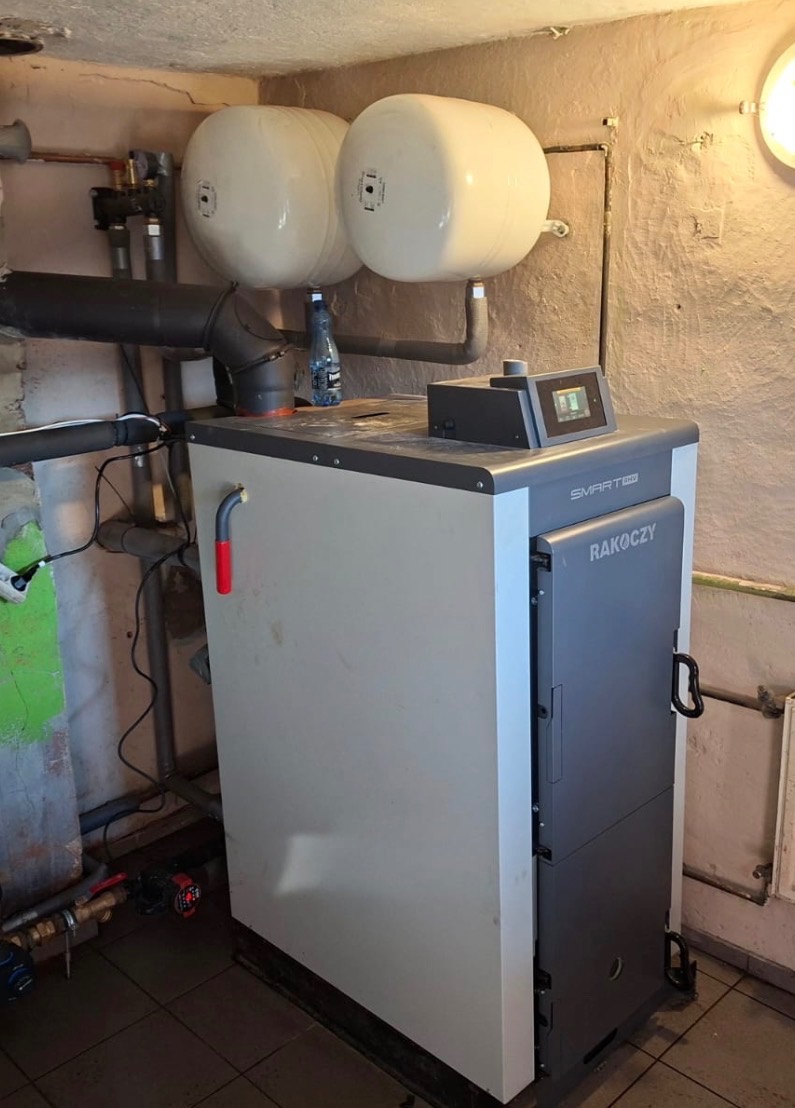 Kocioł grzewczy Rakoczy z dwoma zbiornikami buforowymi w kotłowni o surowych ścianach. Widoczne rury i armatura. Ujęcie z bliska, prezentujące zainstalowane urządzenie.