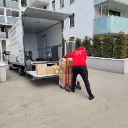 Pracownik w czerwonej bluzie z napisem firmy transportowej przewozi wózkiem kartony do otwartej paki białego busa na tle bloku mieszkalnego z przeszklonymi balkonami.