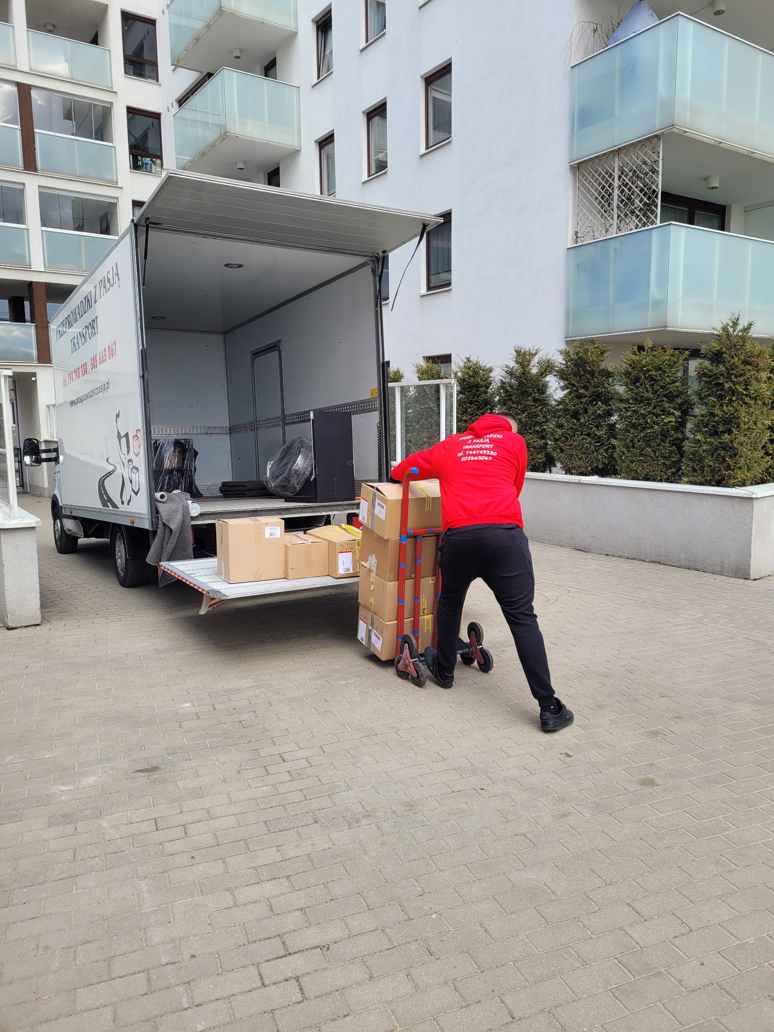 Pracownik w czerwonej bluzie z napisem firmy transportowej przewozi wózkiem kartony do otwartej paki białego busa na tle bloku mieszkalnego z przeszklonymi balkonami.
