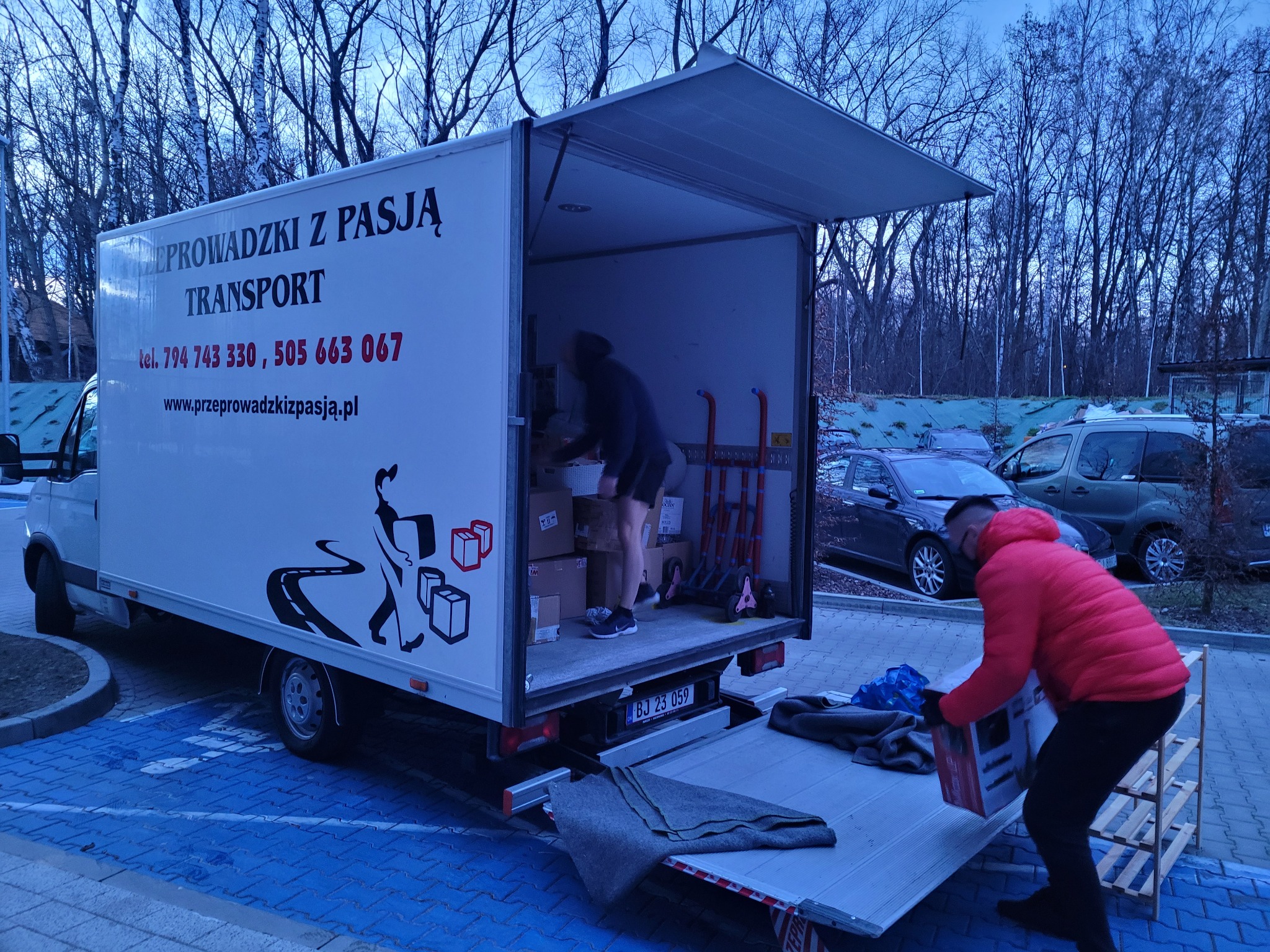 Otwarty samochód dostawczy z napisem 'Przeprowadzki z pasją', pracownik w czerwonej kurtce przenosi karton z samochodu na rampę, drugi pracownik układa kartony wewnątrz, w tle drzewa i zaparkowane...