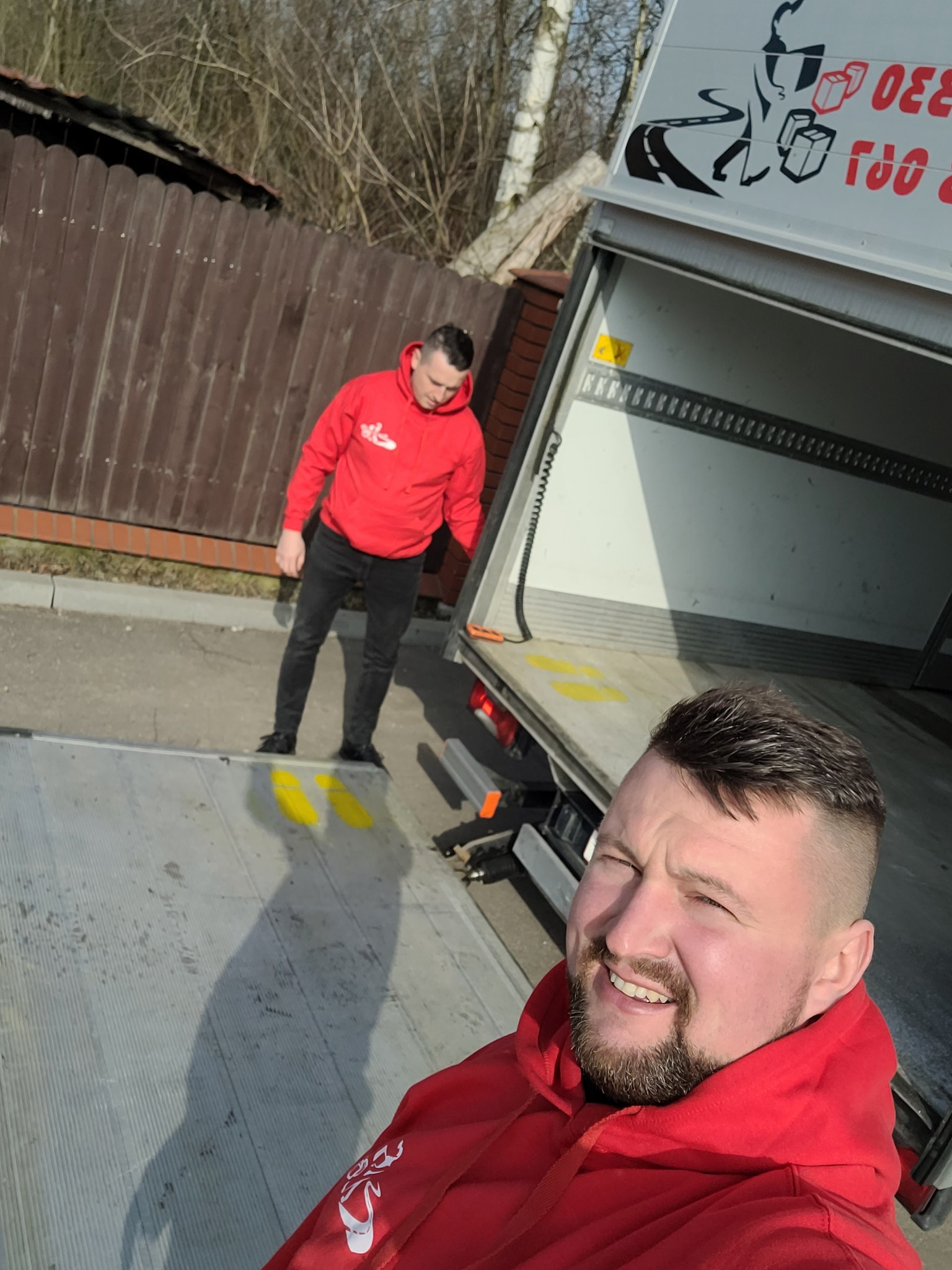 Mężczyzna robiący selfie na rampie auta dostawczego z otwartymi drzwiami, w tle drugi mężczyzna w czerwonej bluzie i brązowe ogrodzenie.