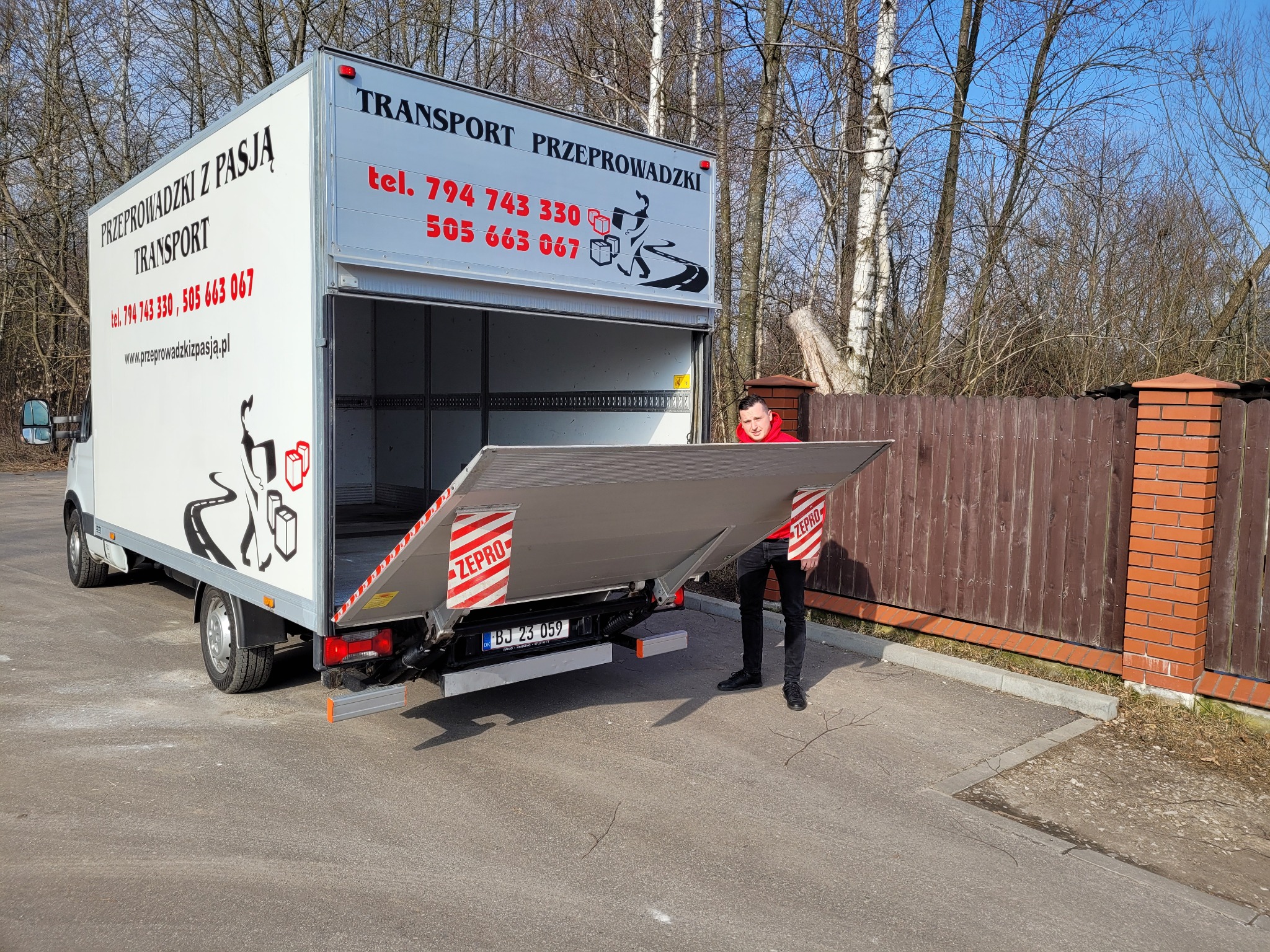 Samochód dostawczy z otwartą windą załadowczą, z widocznym mężczyzną w czerwonej bluzie, na boku nadruk z nazwą firmy i numerami telefonów, w tle drzewa i płot.