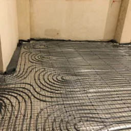 Rury ogrzewania podłogowego rozłożone na folii izolacyjnej i siatce w surowym wnętrzu budynku, widoczne fragmenty ścian i kolumna.