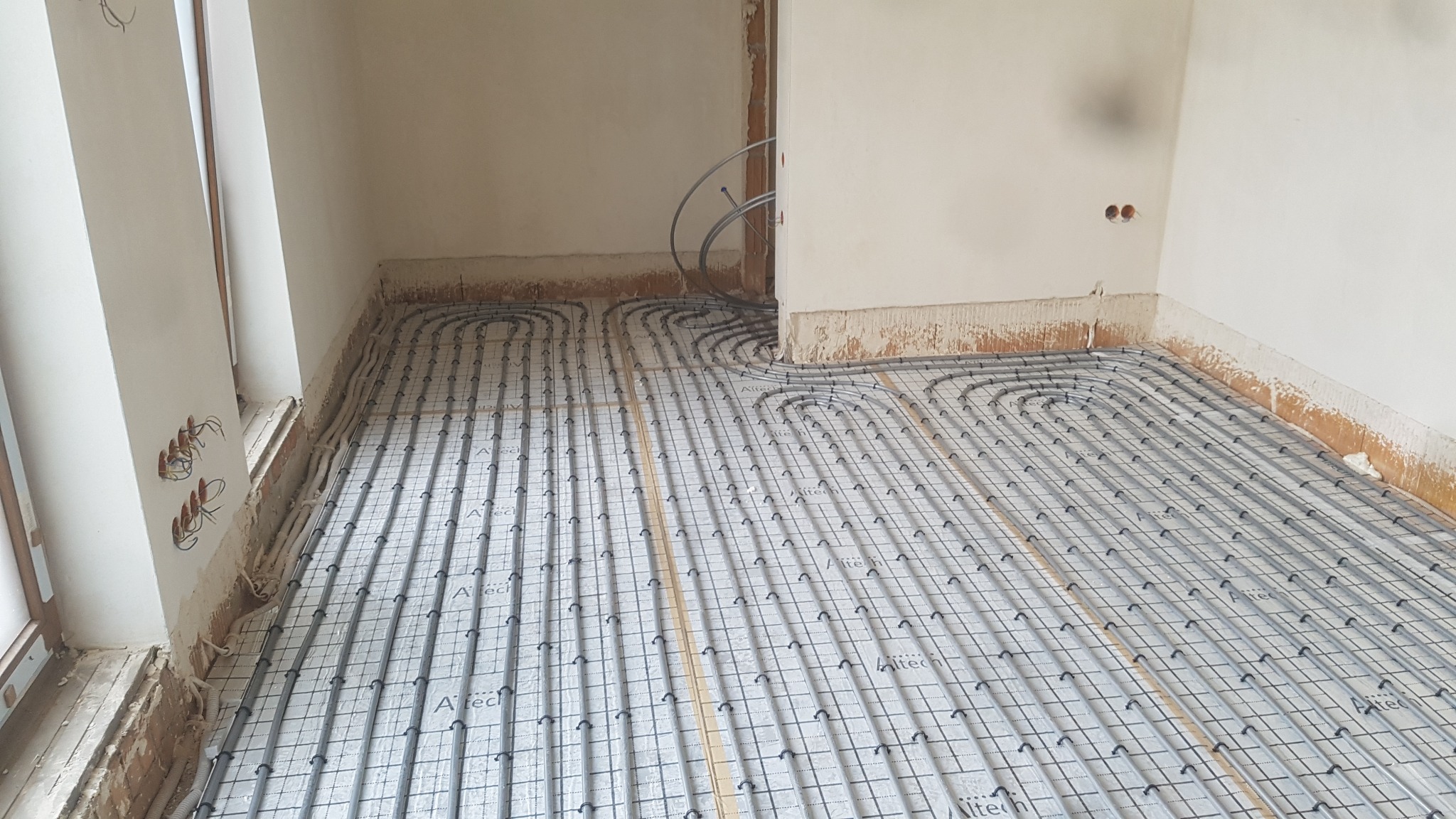 Instalacja ogrzewania podłogowego w trakcie montażu, widoczne rury grzewcze ułożone na izolacji z siatką, surowe ściany w tle, przygotowane punkty elektryczne.