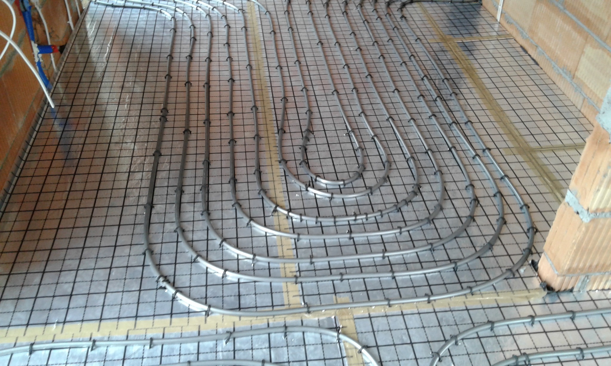 Szare rury ogrzewania podłogowego ułożone w pętle na metalowej siatce, przymocowane czarnymi klipsami, widoczne fragmenty ścian z cegły dziurawki.