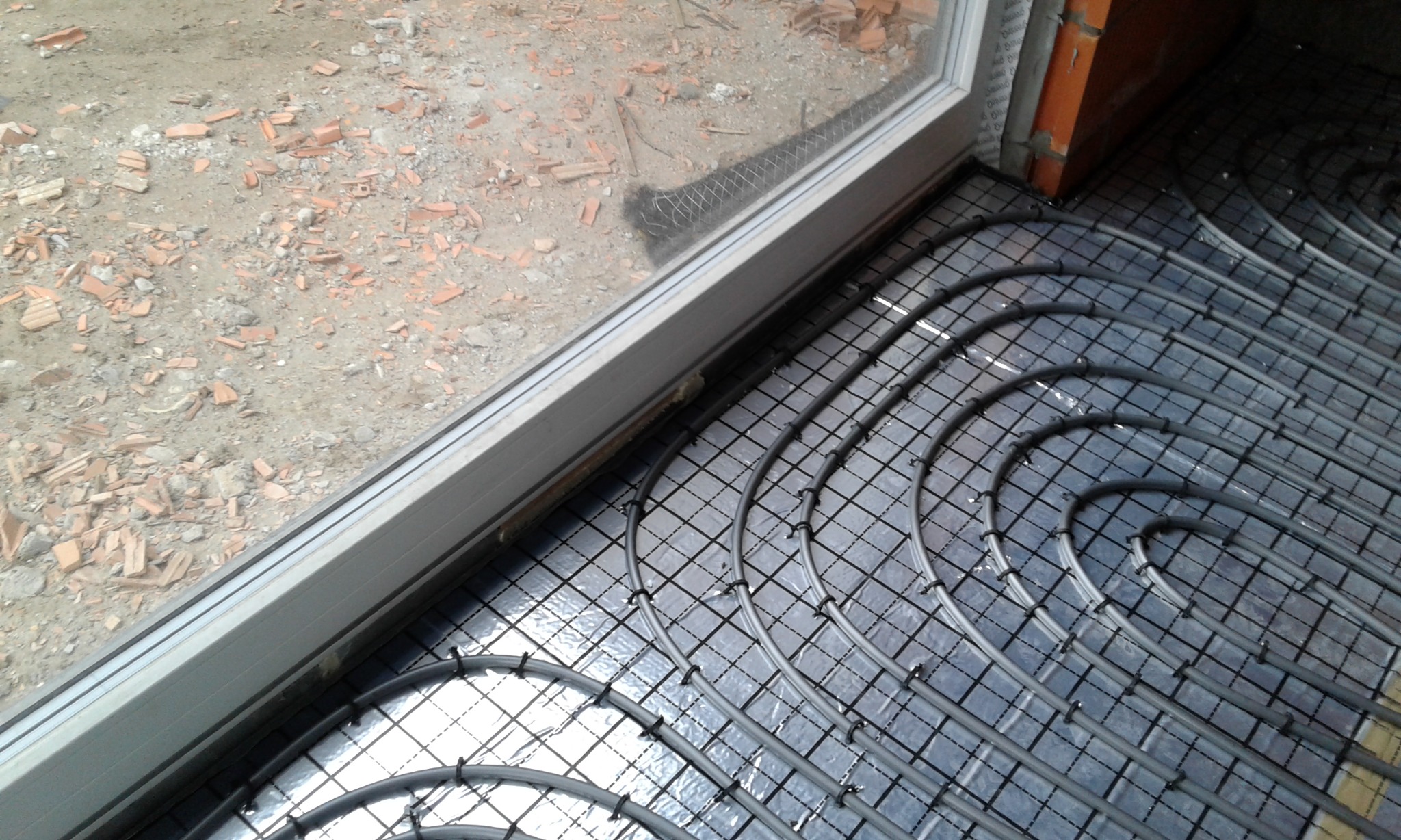 Szare rury ogrzewania podłogowego ułożone w pętle na czarnej macie z siatką, widok z góry, fragment okna i gruz na zewnątrz.