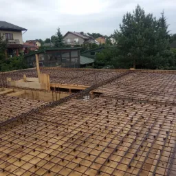 Zbrojenie płyty fundamentowej z drewnianym szalunkiem na tle zabudowy jednorodzinnej w pochmurny dzień.