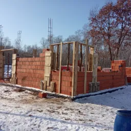 Budowa domu w stanie surowym otwartym: ściany z czerwonej cegły, drewniane szalunki, zbrojenia, zimowa sceneria z pokrywą śnieżną.