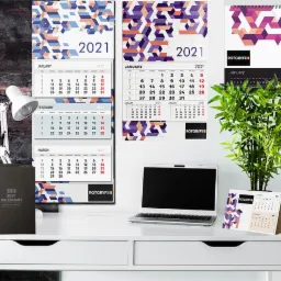 Prezentacja kalendarzy firmowych: ścienne, biurkowe i książkowe, z logo firmy, na tle biurka z laptopem i rośliną doniczkową.