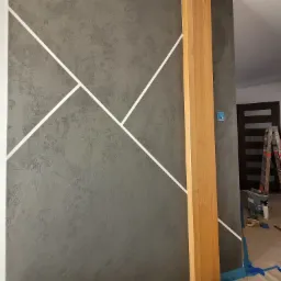 Szara ściana z dekoracyjną fakturą i białymi, geometrycznymi liniami, drewniany filar, podłoga zabezpieczona taśmą malarską, drabina w tle.