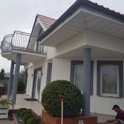 MALINIAK.pl - Elewacja domu jednorodzinnego po remoncie, z balkonem i widoczną osobą układającą płytki na zewnątrz budynku.