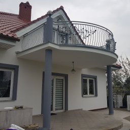 MALINIAK.pl - Biały dom z czerwonym dachem i balkonem z ozdobną, metalową balustradą w kształcie gałązek z liśćmi; widoczne okna z szarymi ramami i drzwi przesuwne.