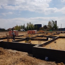 MALINIAK.pl - Fundamenty domu jednorodzinnego w trakcie budowy, zabezpieczone czarną izolacją, zbrojenie stalowe i palety cegieł w tle, pracownik oparty o łopatę.