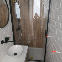 MALINIAK.pl - Nowoczesna łazienka z kabiną prysznicową typu walk-in z czarnymi profilami, umywalką nablatową na białym blacie, szarą cegiełkową glazurą i drewnopodobną okładziną wewnątrz kabiny; widoczne krople...