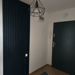 MALINIAK.pl - Nowoczesny przedpokój z czarną ścianą z paneli dekoracyjnych, geometryczną lampą wiszącą, ciemnymi drzwiami wejściowymi i brązową podłogą z płytek.