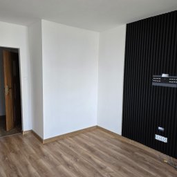 MALINIAK.pl - Pusty pokój po remoncie z panelami ściennymi w kolorze czarnym, drewnianą podłogą i białymi ścianami, widoczny fragment otwartych drzwi.
