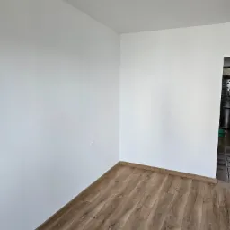 Pusty pokój po remoncie: świeżo pomalowane białe ściany, ułożona podłoga z paneli w odcieniach brązu, widoczne listwy przypodłogowe i gniazdko elektryczne na ścianie, fragment sąsiedniego...