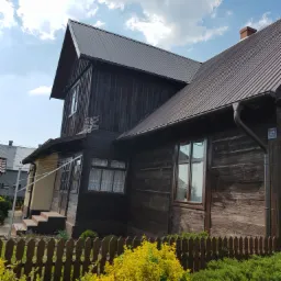 Ciemnobrązowy, drewniany dom z nowym, metalowym dachem w kolorze grafitowym, widoczny numer domu '42' na ścianie, drewniany płot i bujna zieleń w ogrodzie.