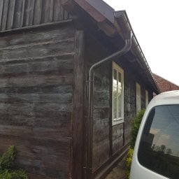 MALINIAK.pl - Odnowiona elewacja drewnianego domu z nowymi, brązowymi rynnami i pomalowaną dolną partią ściany; widoczny fragment białego samochodu.