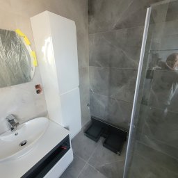 MALINIAK.pl - Łazienka w trakcie wykańczania: szare płytki imitujące marmur, biała szafka łazienkowa z umywalką, kabina prysznicowa z widocznym odbiciem fotografa i zabezpieczone folią lustro z żółtą taśmą.