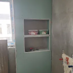 Ściana z płyt kartonowo-gipsowych z wnękami, w których znajdują się pojemniki z materiałami budowlanymi i kielnia, widoczne instalacje wodne na surowej ścianie obok okna.