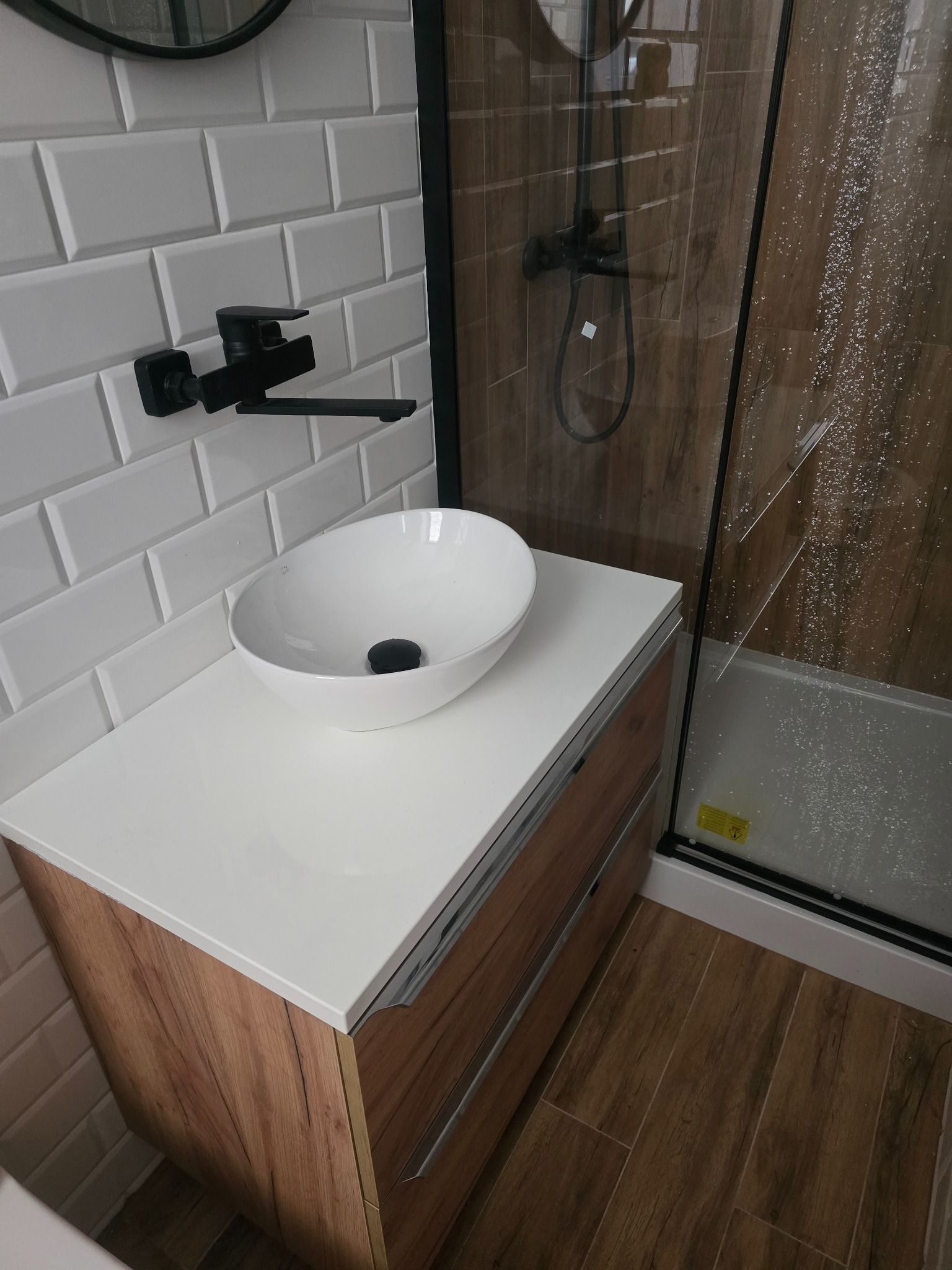 Nowoczesna łazienka w Kaliszu po remoncie: biała umywalka nablatowa z czarną armaturą na drewnianej szafce, białe płytki imitujące cegłę na ścianie i kabina prysznicowa z efektem deszczu.