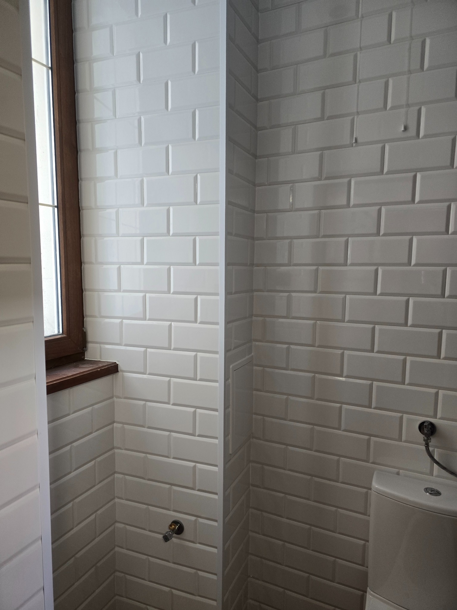 Ściany łazienki wyłożone białymi, prostokątnymi płytkami przypominającymi cegiełki z widocznym oknem z brązową ramą i fragmentem toalety.