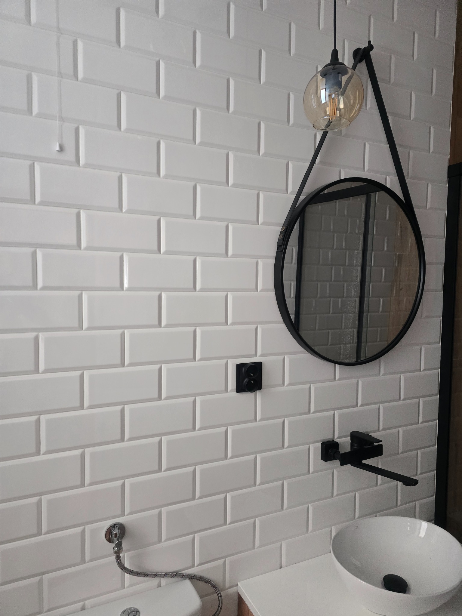 Białe, prostokątne płytki imitujące cegłę na ścianie łazienki, z czarną armaturą, okrągłym lustrem na pasku i szklaną lampą wiszącą, tworzące nowoczesny wystrój.