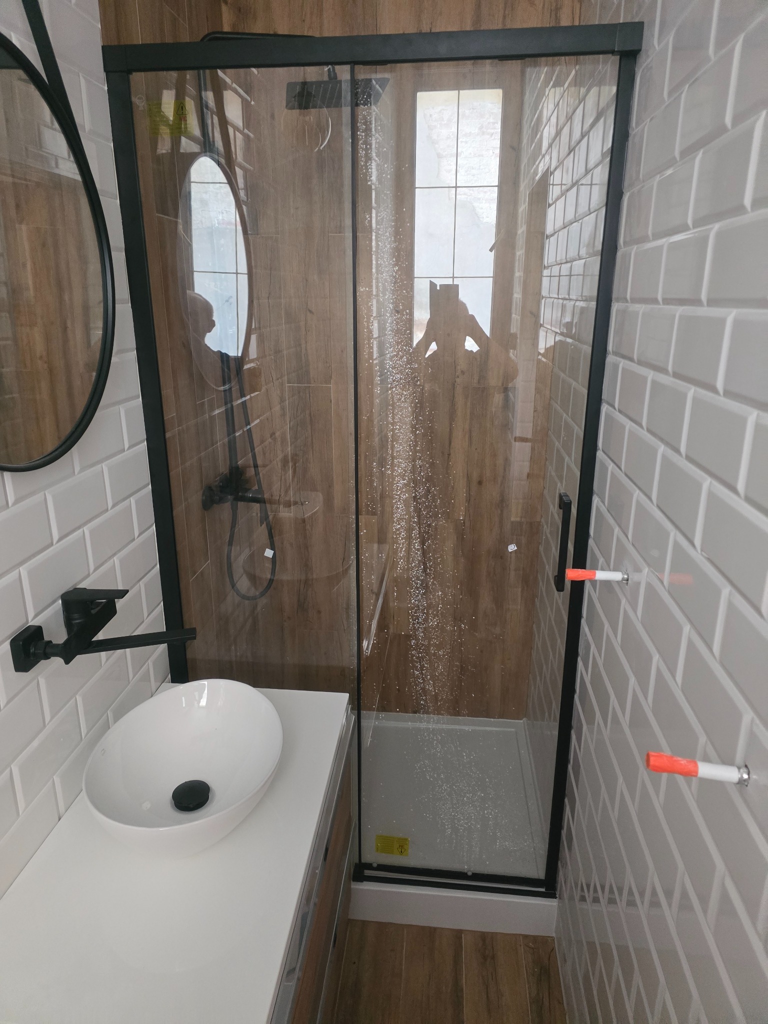 Nowoczesna łazienka z kabiną prysznicową typu walk-in z czarnymi profilami, umywalką nablatową na białym blacie, szarą cegiełkową glazurą i drewnopodobną okładziną wewnątrz kabiny; widoczne krople...