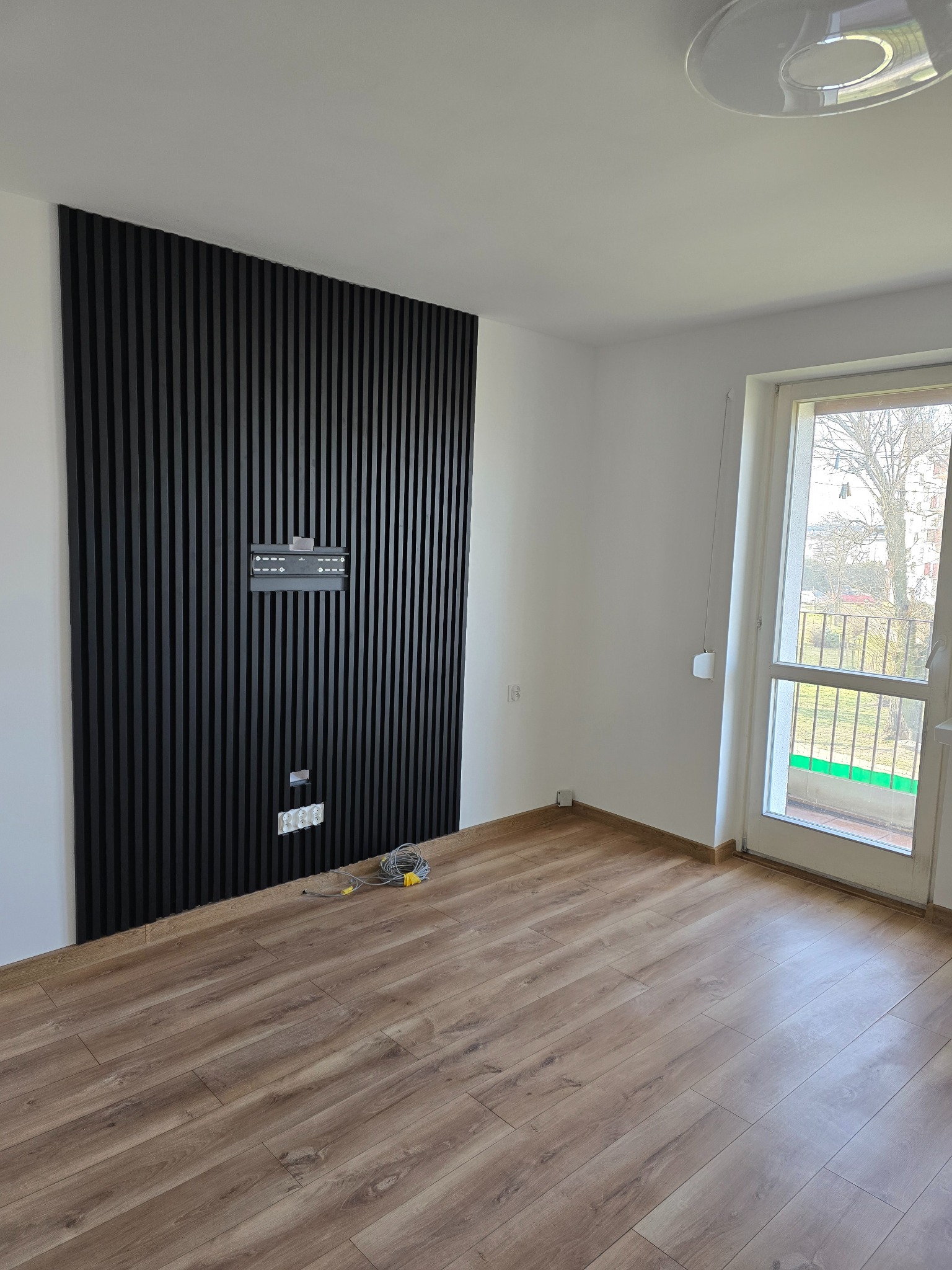 Pokój z panelami ściennymi z czarnych listew, przygotowany do montażu telewizora, widoczne gniazdka elektryczne i przewody.