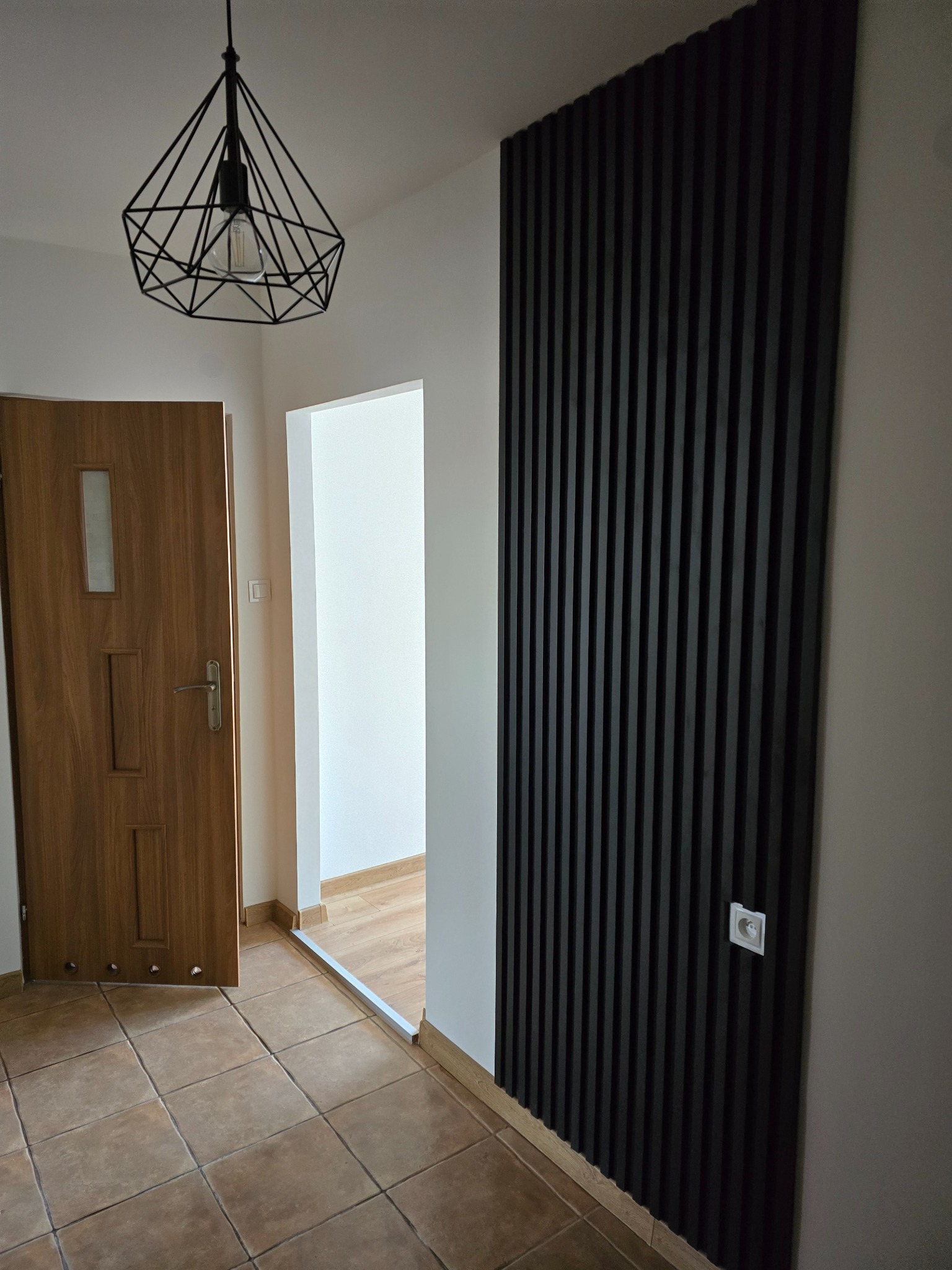 Nowoczesny przedpokój z drewnianymi drzwiami, płytkami podłogowymi i czarnymi panelami ściennymi z włącznikiem światła, oświetlony geometryczną lampą wiszącą.