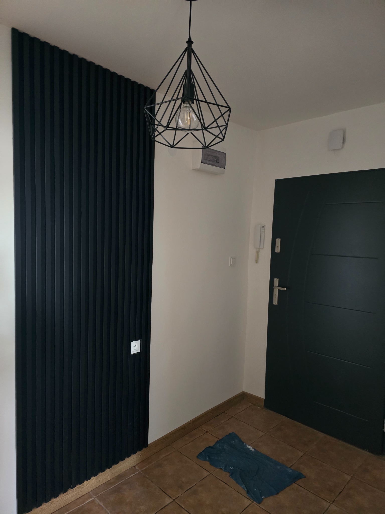 Nowoczesny przedpokój z czarną ścianą z paneli dekoracyjnych, geometryczną lampą wiszącą, ciemnymi drzwiami wejściowymi i brązową podłogą z płytek.