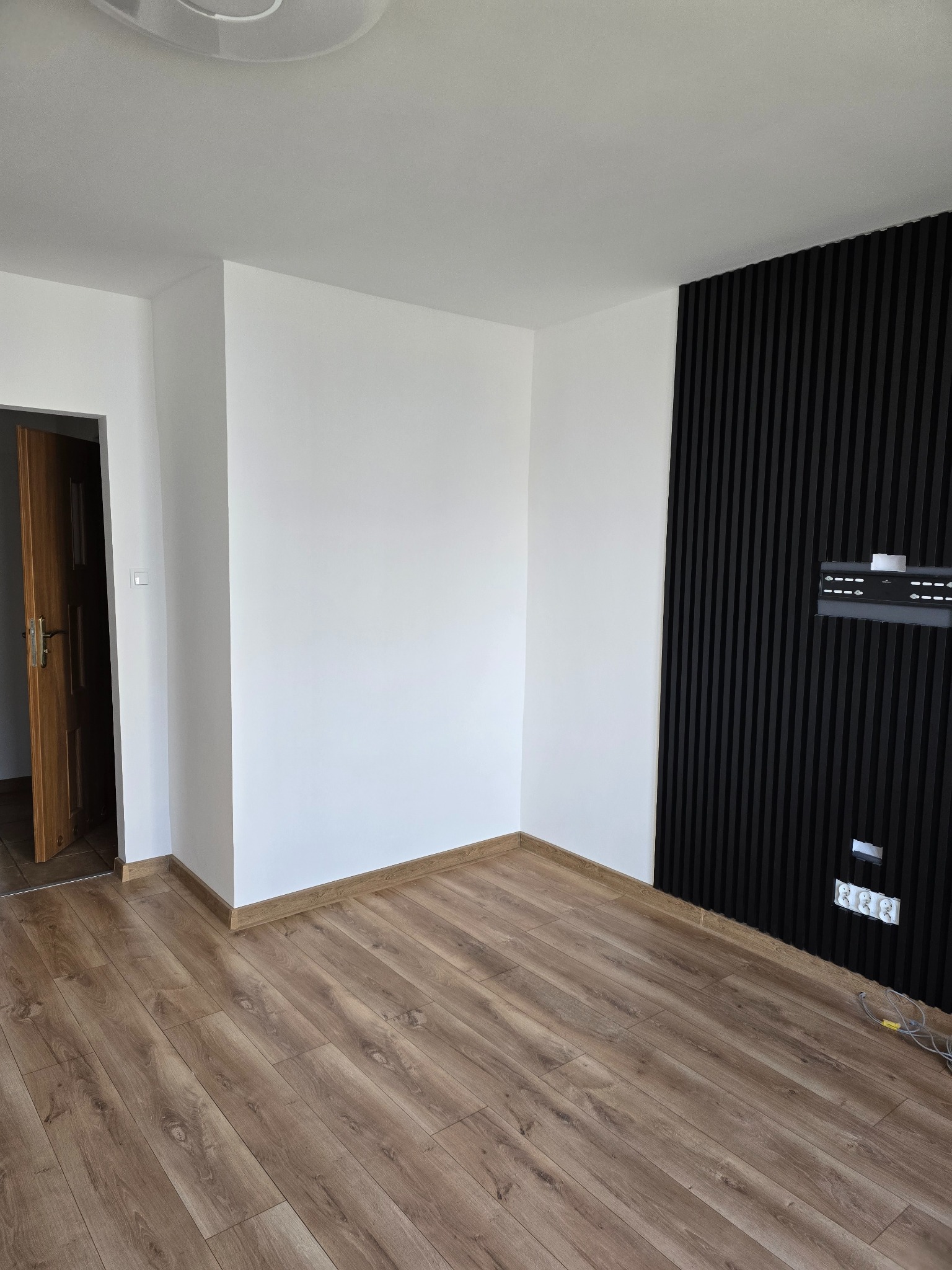 Pusty pokój po remoncie z panelami ściennymi w kolorze czarnym, drewnianą podłogą i białymi ścianami, widoczny fragment otwartych drzwi.