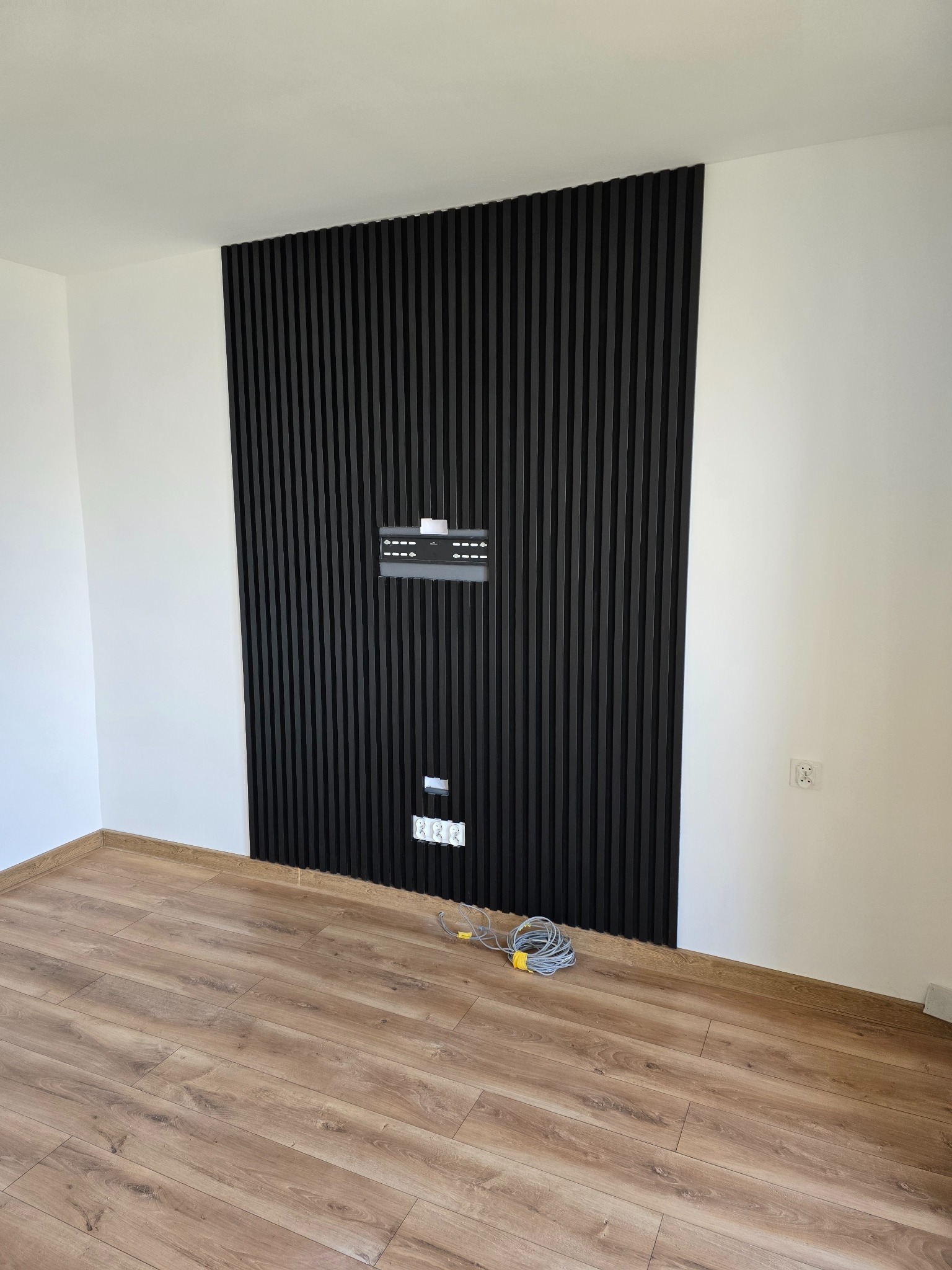 Ściana z czarnych paneli dekoracyjnych z zamontowanym uchwytem na telewizor i gniazdkami elektrycznymi, widoczne kable z żółtą opaską na podłodze z paneli.