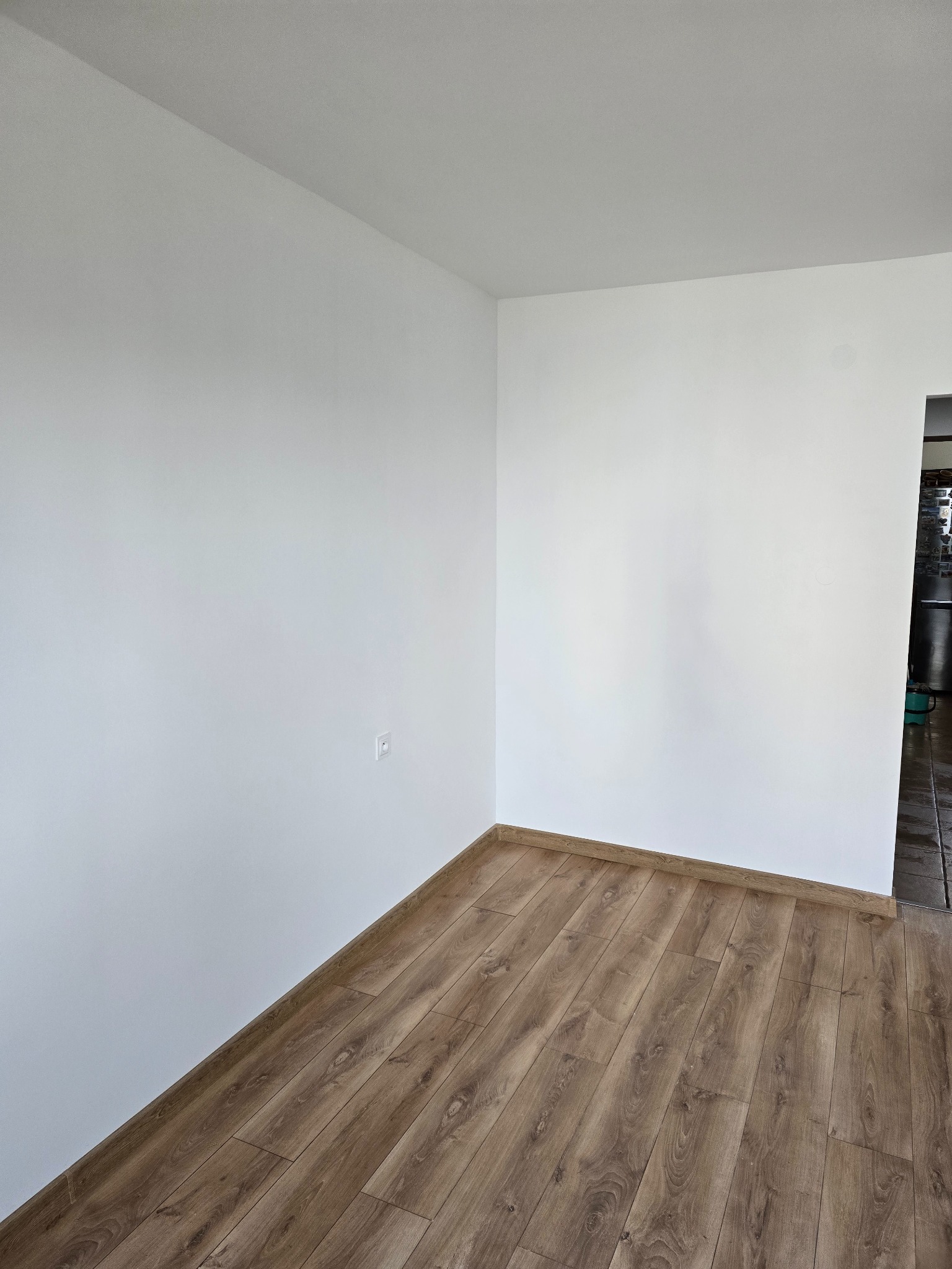 Pusty pokój po remoncie: świeżo pomalowane białe ściany, ułożona podłoga z paneli w odcieniach brązu, widoczne listwy przypodłogowe i gniazdko elektryczne na ścianie, fragment sąsiedniego...