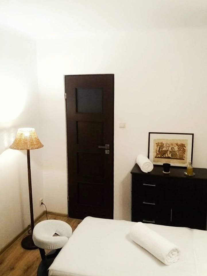 Jasne wnętrze gabinetu z łóżkiem do zabiegów, stolikiem z ręcznikami i obrazkiem, oraz lampą z wiklinowym abażurem, ukazane w minimalistycznym stylu.