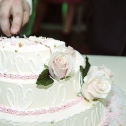 śluby foto-wideo - Krojenie trzypiętrowego tortu weselnego z białym lukrem, różowymi zdobieniami i świeżymi różami, widoczna dłoń w garniturze z obrączką.