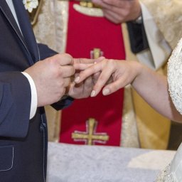 śluby foto-wideo - Moment zakładania obrączki podczas ceremonii ślubnej, widoczny fragment sukni ślubnej z koronkowymi zdobieniami i dłonie nowożeńców na tle szaty liturgicznej.