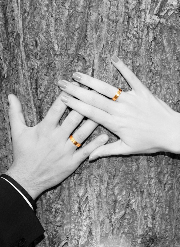 Dłonie pary młodej z złotymi obrączkami na tle kory drzewa, zdjęcie czarno-białe z wyeksponowanymi obrączkami w kolorze złotym, widoczny fragment rękawa marynarki pana młodego.