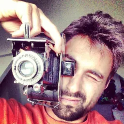 Mężczyzna z przymrużonym okiem trzyma przed twarzą starodawny aparat fotograficzny, widoczne detale obiektywu i metalowej obudowy.