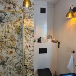 Nowoczesna łazienka z kabiną prysznicową z motywem kwiatowym i ptaków, czarna toaleta i umywalka, oświetlenie w stylu industrialnym z elementami złota, białe płytki na ścianach.